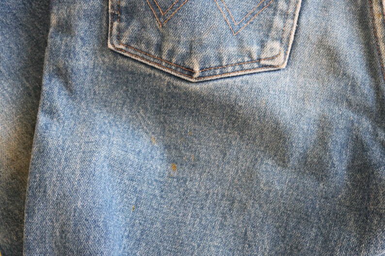 May include: Close-up of blue denim fabric with a back pocket. The fabric has a worn, faded look with visible stitching and a slight sheen.