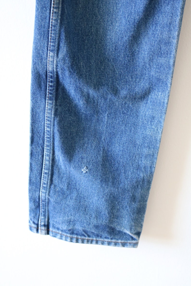 May include: Close-up of the bottom of a pair of blue denim jeans. The jeans have a single seam down the side and a small hole in the fabric.