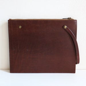 May include: A brown leather clutch with a zipper closure and a short strap. The clutch has a subtle textured finish.