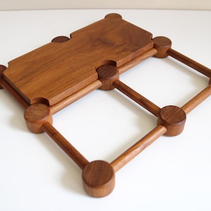 May include: A wooden serving tray with a geometric design. The tray is made of a warm-toned wood and features a rectangular top section supported by a diamond-shaped frame. The frame is constructed with rounded wooden posts and connecting rods.