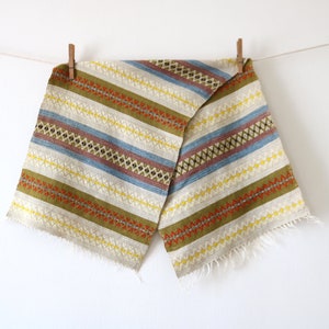 May include: A woven fabric table runner with a fringed edge. The runner features a striped pattern in shades of green, yellow, blue, and red.
