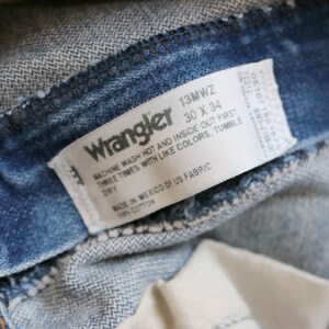 May include: A white Wrangler jeans care label with the text "13MWZ 30 X 34" and instructions for washing and drying the jeans. The label is attached to a pair of blue denim jeans.