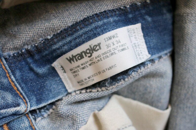 May include: A white Wrangler jeans care label with the text "13MWZ 30 X 34" and instructions for washing and drying the jeans. The label is attached to a pair of blue denim jeans.