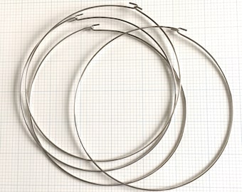 May include: Four silver-colored metal embroidery hoops of varying sizes are arranged on a grid background. Each hoop has a small clasp at the top. The hoops are circular and appear to be made of thin wire.
