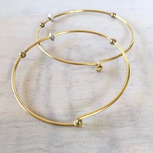 Può includere: Due braccialetti rigidi dorati con piccole perle rotonde. I braccialetti sono aperti e hanno una chiusura a molla.