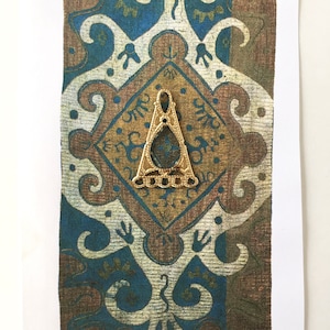 May include: A gold-colored metal pendant in the shape of a triangle with intricate details. The pendant is set against a background of a patterned fabric with blue and brown colors.