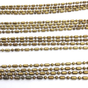 May include: Multiple strands of gold-toned metal chain with round and elongated links. The chain is laid out in parallel lines on a white surface.