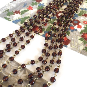 May include: Close-up of several strands of dark brown beaded necklaces with gold-coloured chain links. The necklaces are layered on a patterned background with floral and geometric designs.