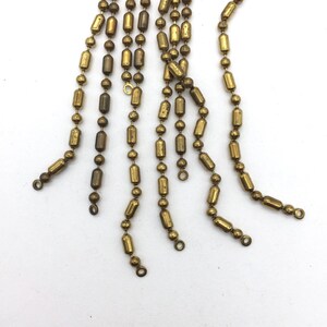 May include: A set of gold-toned metal chains with cylindrical beads. The chains are all different lengths and are hanging down from a single point.