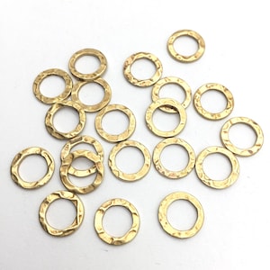 May include: A collection of small, hammered gold-colored metal rings. The rings have a textured surface and a circular shape with a hole in the center. They are scattered on a white surface.