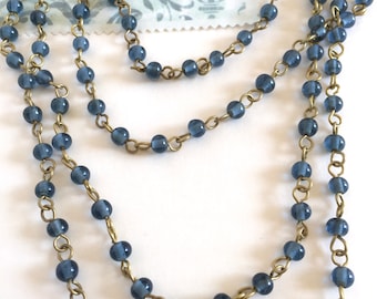 May include: A strand of blue glass beads strung on a brass chain. The beads are round and have a slightly frosted finish.