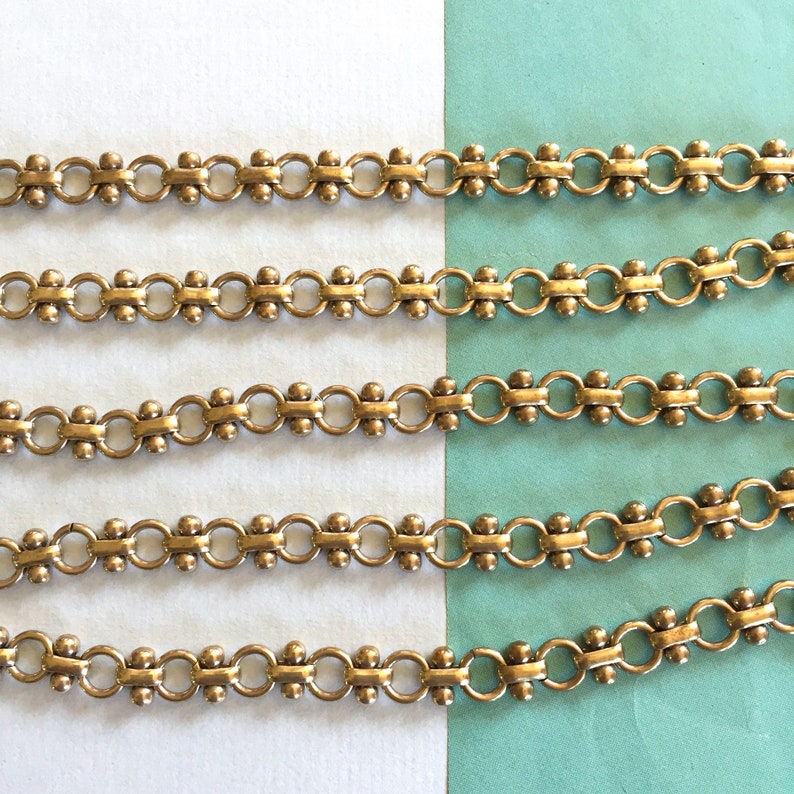 May include: Five rows of gold-toned chain with round links and small beads. The chain is on a white and blue background.