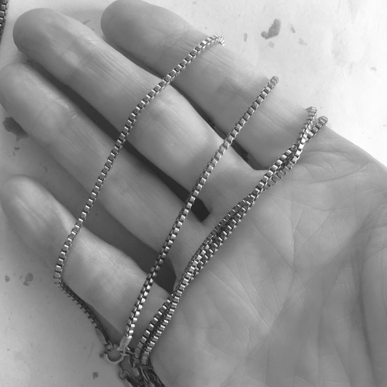 May include: A silver chain necklace with a box chain design. The chain is folded in half and held in a hand.