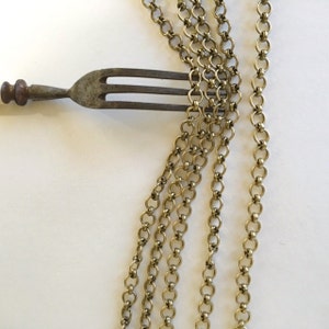 May include: A close-up of four strands of gold chain, each with a different length. The chains are made of small, round links. A fork is lying on top of the chains.