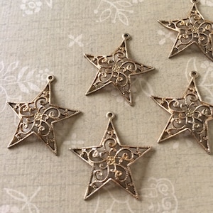 May include: Five gold-colored star charms with intricate filigree designs. Each star has a small loop at the top for hanging. The stars are arranged on a light-colored patterned surface.
