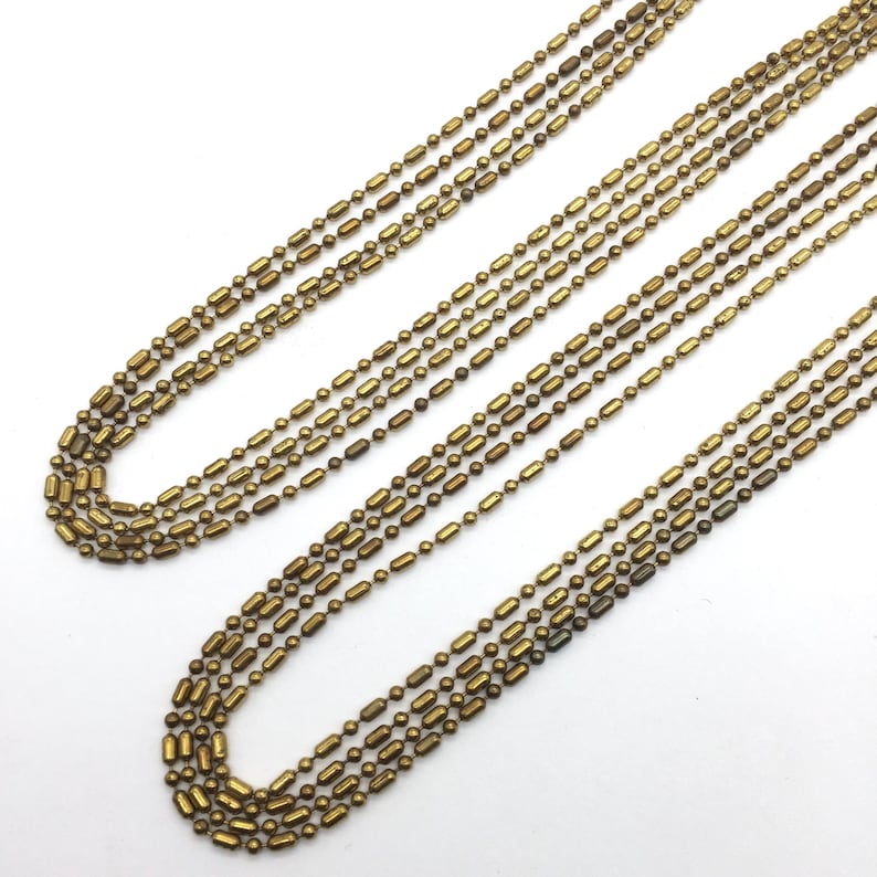 May include: A set of six gold-toned metal chains with a cylindrical link design. The chains are arranged in a layered pattern, with the longest chain at the top and the shortest chain at the bottom.