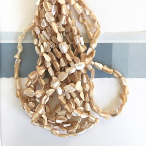 May include: A strand of beige shell beads with an iridescent finish. The beads are irregularly shaped and have a natural, organic look.