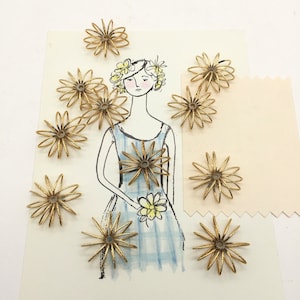 May include: A set of 12 gold metal flower embellishments. The embellishments are delicate and have a wire-like construction. They are arranged on a white background with a drawing of a woman in a blue and white dress.