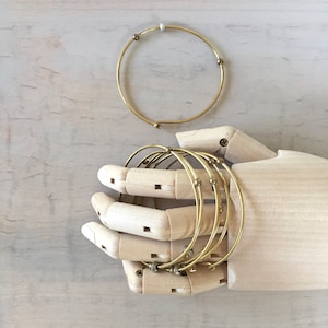 May include: A set of six gold-toned bangle bracelets with small beads on each bracelet. The bracelets are stacked on a wooden hand model.