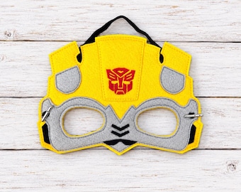 May include: A yellow and gray felt mask featuring a red Autobot symbol in the center. The mask has two eye openings, black accents, and a black elastic strap attached to the top for securing the item around the head during use.
