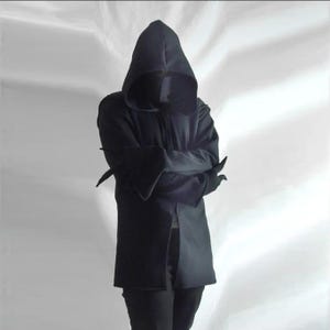 Könnte beinhalten: Eine schwarze Kapuzenjacke mit langen Ärmeln, ausgestellten Bündchen und einem durchgehenden Frontreißverschluss. Das Kleidungsstück besteht aus einfarbigem schwarzem Stoff und verfügt über eine tiefe, spitz zulaufende Kapuze, die den Oberkörper verdeckt.