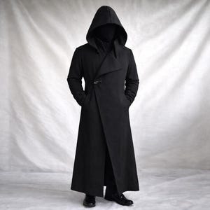 May include: A long black coat featuring a deep hood, a high collar, and a single metallic toggle closure at the chest. The garment has long sleeves, a straight hemline, and a structured silhouette that extends to the lower calf area.