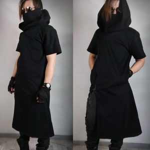 May include: A black hooded tunic with a long, flowing skirt and short sleeves. The tunic has a high collar and a zipper down the front. The model is wearing black pants and black boots.
