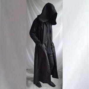 Peut inclure: Un long manteau noir à capuche doté d'une capuche structurée, de manches longues et d'une silhouette pleine longueur. Le manteau est porté sur une chemise noire et un pantalon noir, créant une apparence monochrome aux lignes épurées et lisses.