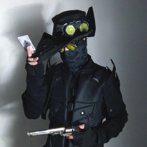 May include: A person wearing a black tactical vest and a black hat with green goggles. They are holding a playing card and a silver pistol.