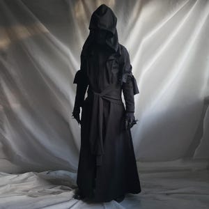 May include: A full-length black hooded robe featuring long sleeves with tied cuffs, a structured hood, and a matching fabric belt tied at the waist. The garment is constructed from a solid black fabric and includes dark gloves with pointed accents.