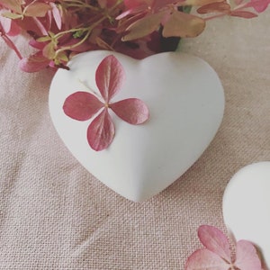 Peut inclure: Un plat en céramique blanc en forme de cœur avec une fleur rose sur le dessus. Le plat est posé sur une surface en tissu beige.