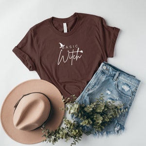 May include: A brown t-shirt with the text "Basic Witch" in white lettering. The shirt is styled with a brown hat, denim shorts, and greenery.
