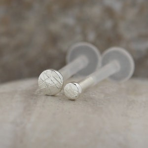 Op de afbeelding: Twee zilveren lichaamsieraden met een getextureerd rond ontwerp. De sieraden bevinden zich op een doorzichtige plastic achterkant.