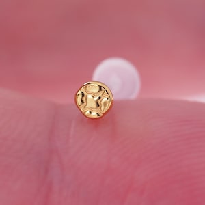 May include: A small, gold-colored stud earring with a textured, circular design. The earring is held against a pink background, showcasing its intricate details and polished finish. The stud's design features a pattern of raised, irregular shapes.