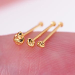 May include: Three gold-colored nose studs with textured designs. The studs are on a finger, with a blurred pink background. The studs have different shapes, with one appearing like a small nugget.