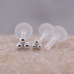 Peut inclure: Deux ensembles de labrets argentés. Chaque labret présente trois petites perles rondes argentées disposées en triangle. Les labrets ont des tiges et des dos transparents et flexibles. Le fond est une surface texturée neutre.