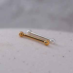 Op de afbeelding: Twee neusringen, een gouden en een zilveren, liggen op een wit oppervlak. De ringen zijn beide recht en hebben aan elk uiteinde kleine ronde balletjes.