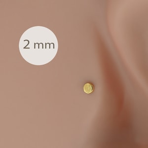 Pode incluir: Um pequeno brinco de botão redondo dourado. O brinco tem 2 mm de diâmetro e é mostrado numa orelha. O brinco tem uma superfície texturizada e um design simples. O fundo é um tom de pele claro.