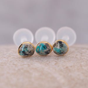 Könnte beinhalten: Drei goldgerahmte Ohrstecker mit einem marmorierten Design aus Türkis, Grau und Schwarz. Die Ohrringe sind auf einer neutralen Oberfläche platziert, wobei die Ohrringverschlüsse leicht unscharf im Hintergrund sind. Die Ohrringe haben eine runde Form.