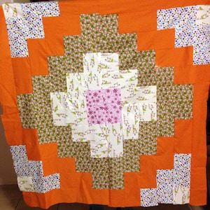 Puede incluir: Una colcha con un patrón geométrico de cuadrados naranjas, verdes, blancos y rosas. La colcha está hecha de diferentes telas con varios patrones.