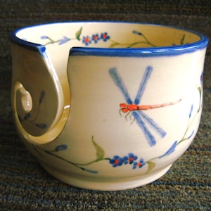 May include: A white ceramic yarn bowl with a blue rim and a dragonfly design. The bowl has a hole in the side for yarn to feed through.