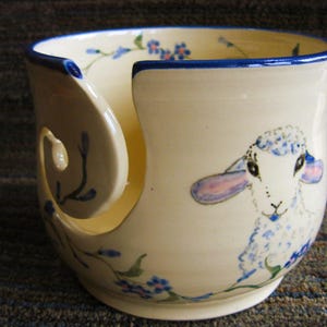 May include: White ceramic yarn bowl with a blue rim and a blue and white floral design. The bowl has a cutout for yarn to feed through and features a painted image of a white lamb with pink ears.
