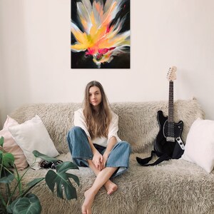 K&ouml;nnte beinhalten: Eine Frau sitzt auf einem flauschigen, hellbraunen Sofa in einem wei&szlig;en Shirt und einer blauen Jeans. Eine schwarze E-Gitarre lehnt am Sofa. Ein abstraktes Gem&auml;lde einer gelben, orangen und pinken Blume auf schwarzem Hintergrund h&auml;ngt an der Wand hinter dem Sofa.