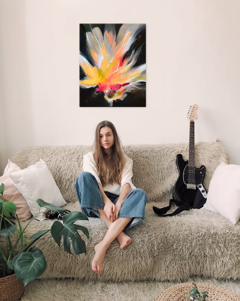 K&ouml;nnte beinhalten: Eine Frau sitzt auf einem flauschigen, hellbraunen Sofa in einem wei&szlig;en Shirt und einer blauen Jeans. Eine schwarze E-Gitarre lehnt am Sofa. Ein abstraktes Gem&auml;lde einer gelben, orangen und pinken Blume auf schwarzem Hintergrund h&auml;ngt an der Wand hinter dem Sofa.