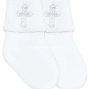 May include: A pair of white baby socks with a folded cuff. Each cuff features a silver cross applique. The text "Accessories by Me" is printed in a light gray color on the socks.