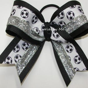 May include: Black and white soccer ball patterned hair bow with silver glitter accents. The bow is tied with a black elastic.