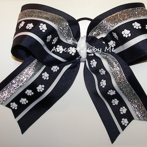 May include: A large navy blue hair bow with white paw prints and silver glitter accents. The bow features a white ribbon and the text "Accessories by Me". This handmade bow is perfect for pet lovers.