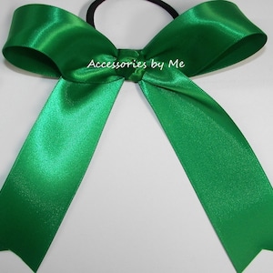 May include: A green satin ribbon bow with a black elastic hair tie. The bow is tied in a classic style with two long tails.