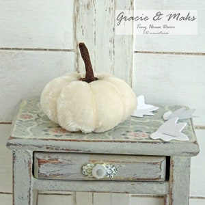 May include: A white velvet pumpkin with a brown stem sits on a small, gray, distressed side table with a drawer. The table has a floral pattern on the top. The text "Gracie & Maks Tiny House Decor 1:12 miniatures" is visible in the background.