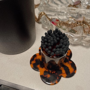May include: A black and brown tortoiseshell flower-shaped dish with a clear glass jar filled with black matches. The jar is sitting on the dish.